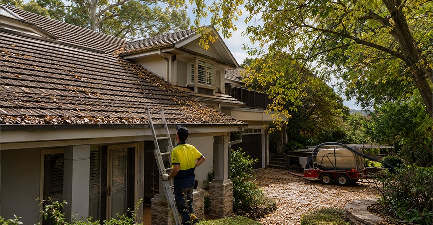 A professional gutter cleaner in high-visibility workwear standing by a ladder, assessing a multi-story house roof heavily covered in dead leaves and debris.